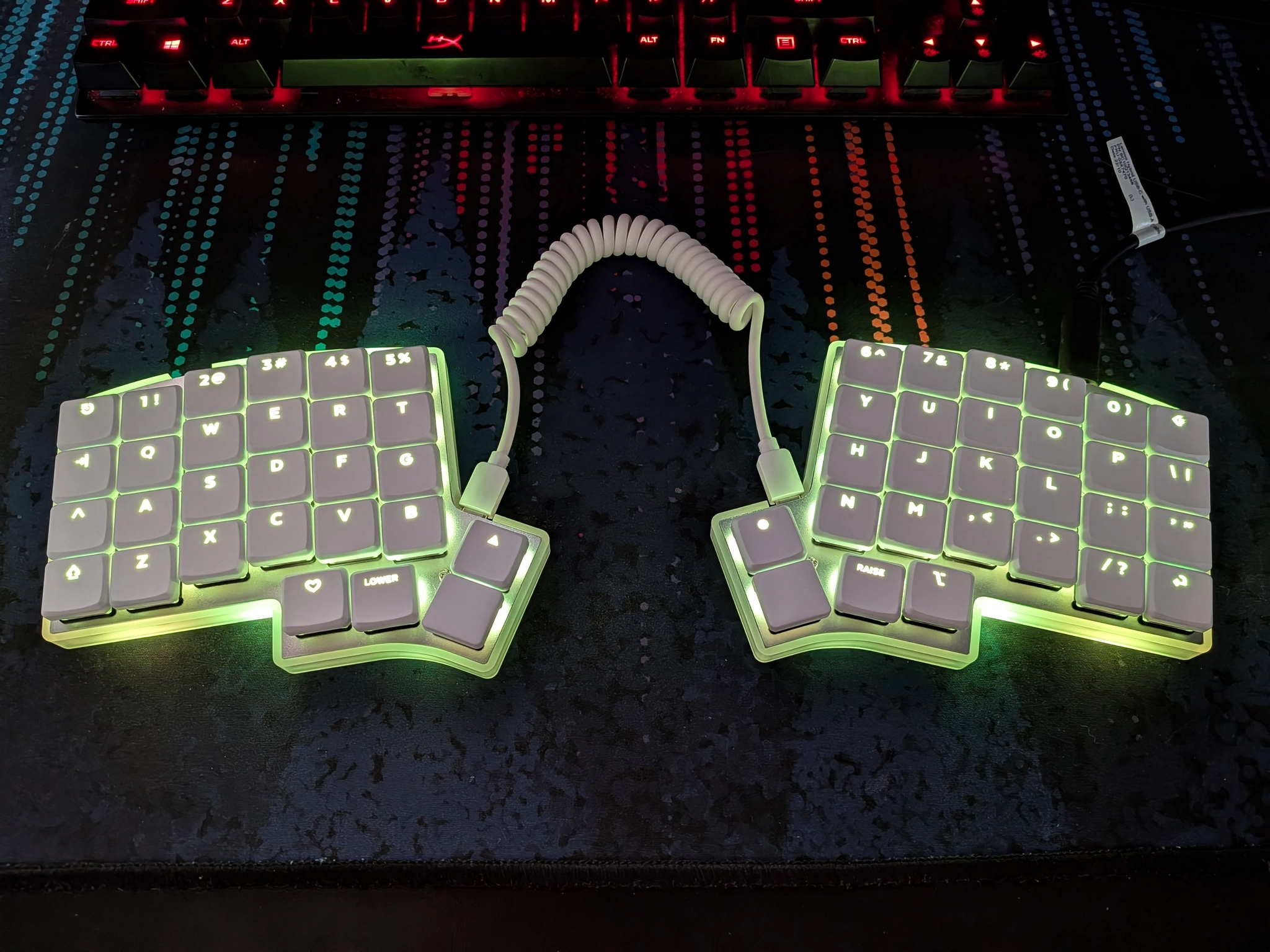 an Iris CE Revision 1 sitting on my desk. it's an ergonomic split (two separate halves) keyboard with addressable RGB backlighting (currently a light green), low-profile keys, and a coiled USB-C cable between the two halves. the whole board is white with light green backlighting, which shines through the keycap legends.

above that is a cropped view of a hyperX alloy fps pro, a black ten-keyless board with red backlighting. even with the crop, it's noticeably bulkier than the new board.